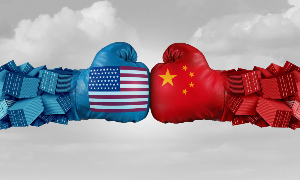  Two boxing gloves, one with an American flag pattern and the other with a Chinese flag pattern, with shipping containers in the background representing the ongoing trade war between the United States and China.